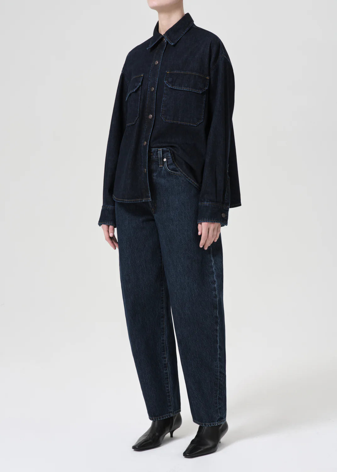 A person wearing a dark denim Gwen Slice Shirt - Rinse by AGOLDE, featuring a classic button-down silhouette and crafted from organic cotton, pairs it with jeans and black shoes while standing against a plain background.