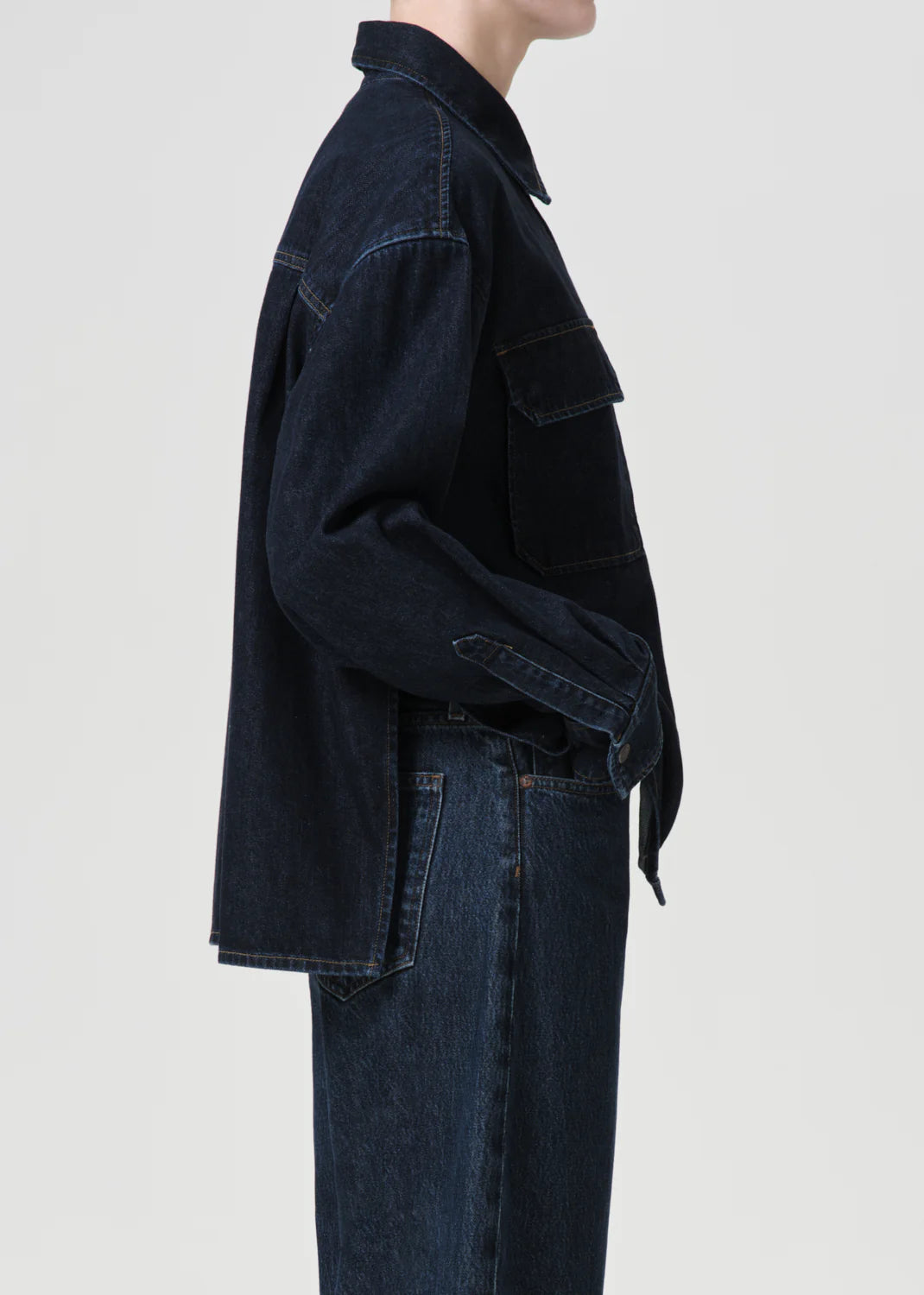 A person in the Gwen Slice Shirt - Rinse by AGOLDE, featuring a dark denim button-down silhouette, paired with jeans and crafted from organic cotton, stands in profile against a plain background.