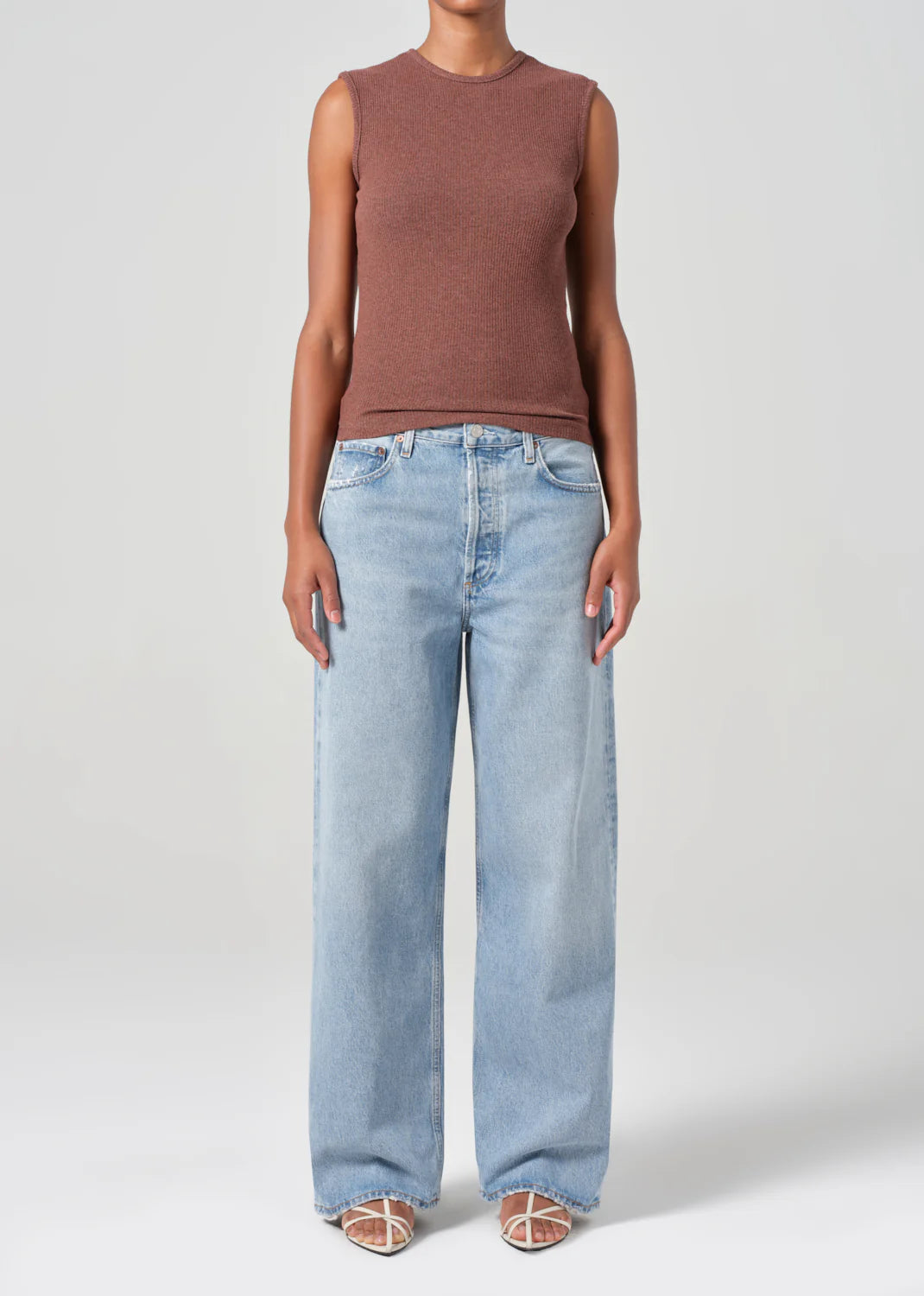 Wearing a sleeveless brown top and AGOLDE's Low Slung Baggy 30.5" jeans in Force, crafted from organic cotton, a person stands against a neutral backdrop. Strappy sandals enhance the effortless style of their oversize ensemble.