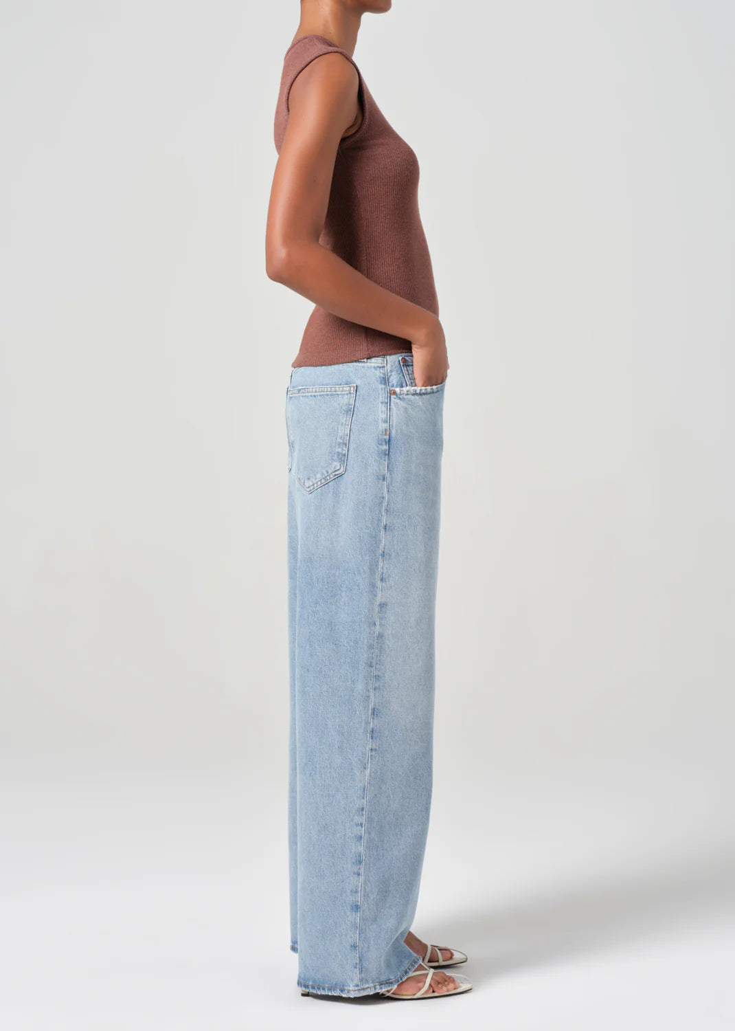 A person in profile wears a sleeveless brown top and light blue AGOLDE Low Slung Baggy 30.5" - Force jeans made from organic cotton. The oversized fit of the jeans effortlessly complements the simple background.