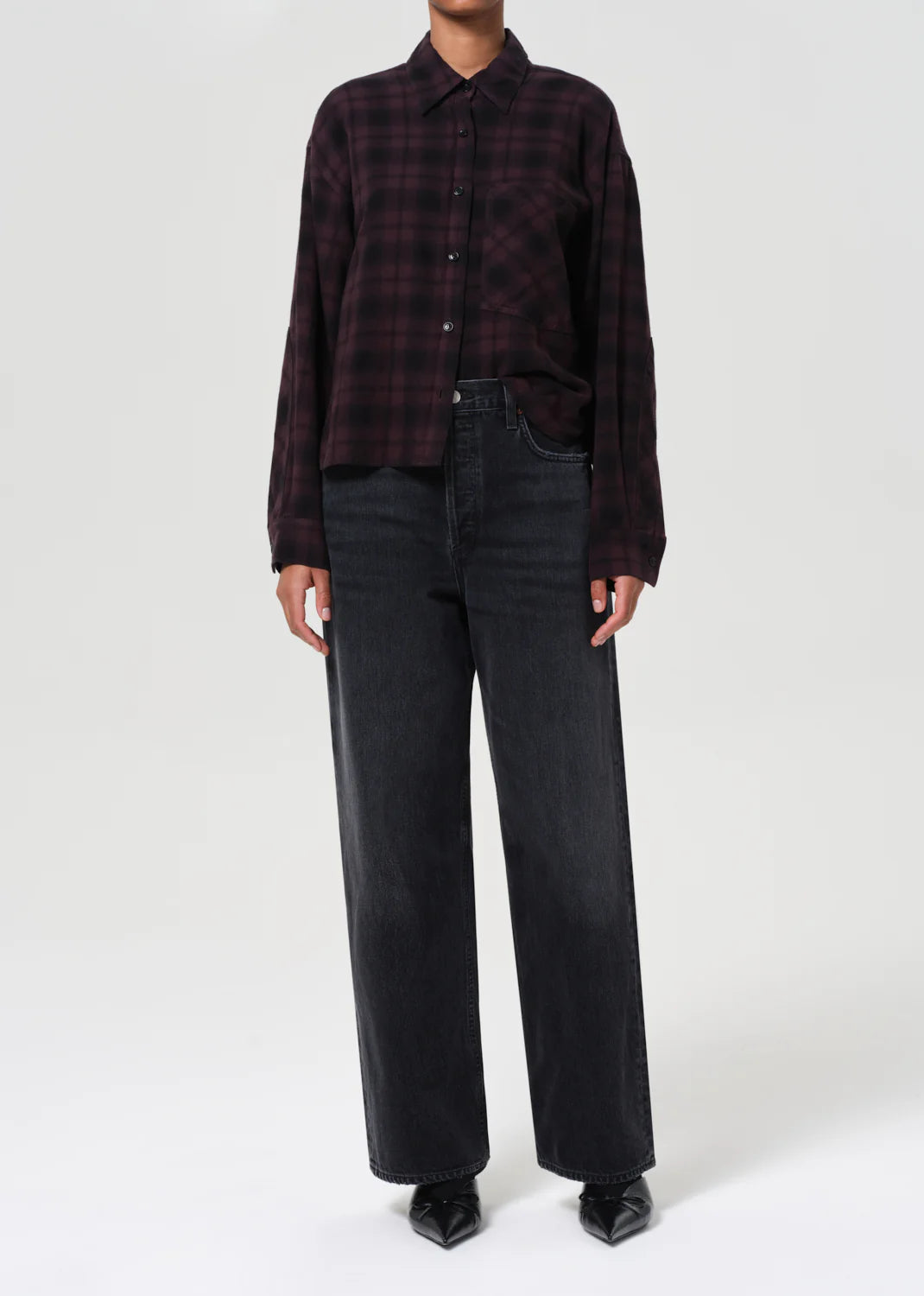 A person stands against a plain light background wearing a dark plaid button-up shirt, AGOLDE’s Low Slung Baggy - Recess black denim jeans, and black pointed shoes.
