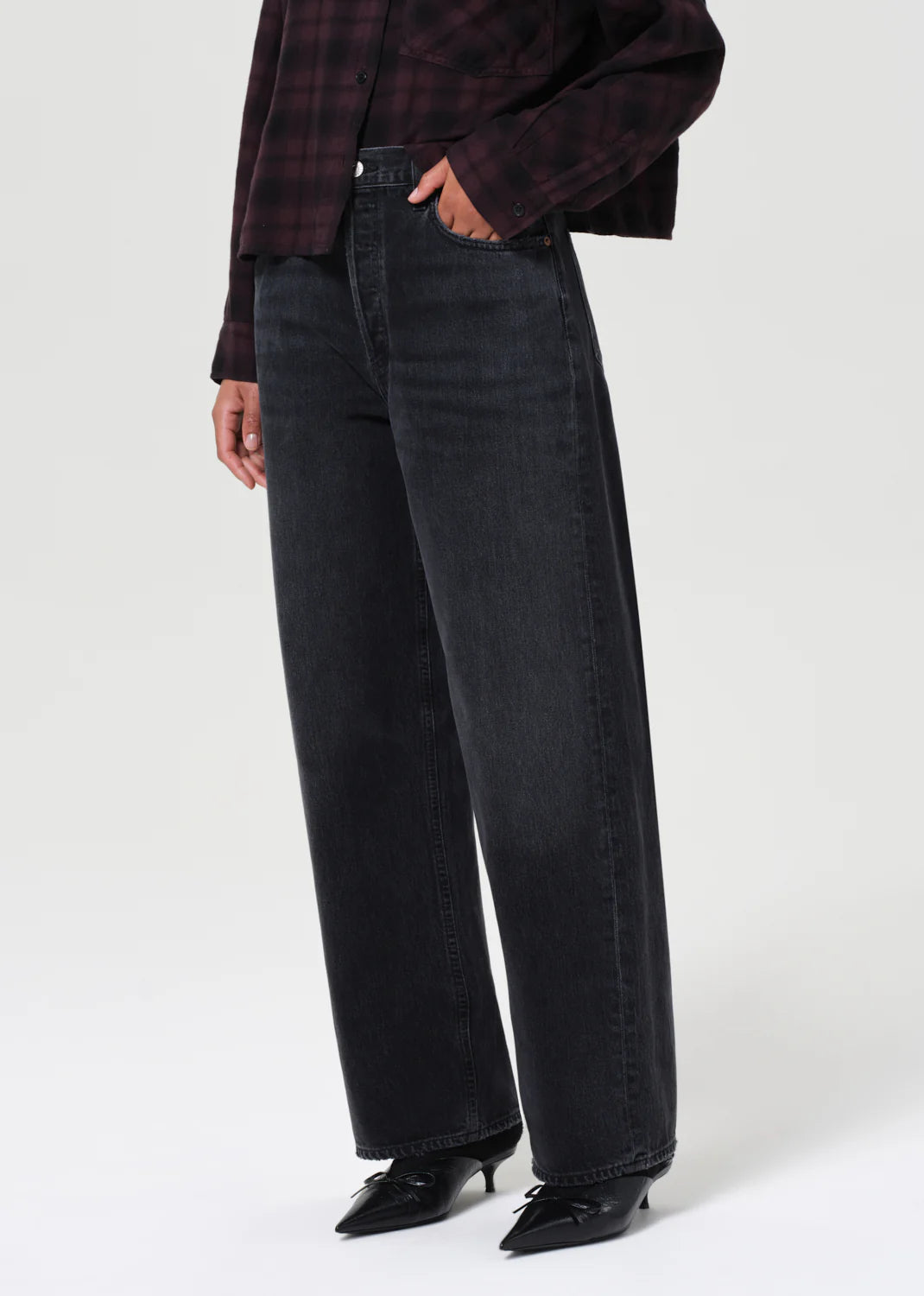A person wears AGOLDE's Low Slung Baggy - Recess black denim jeans made from regenerative cotton, paired with a dark plaid shirt and black pointed-toe shoes, standing against a plain light background.