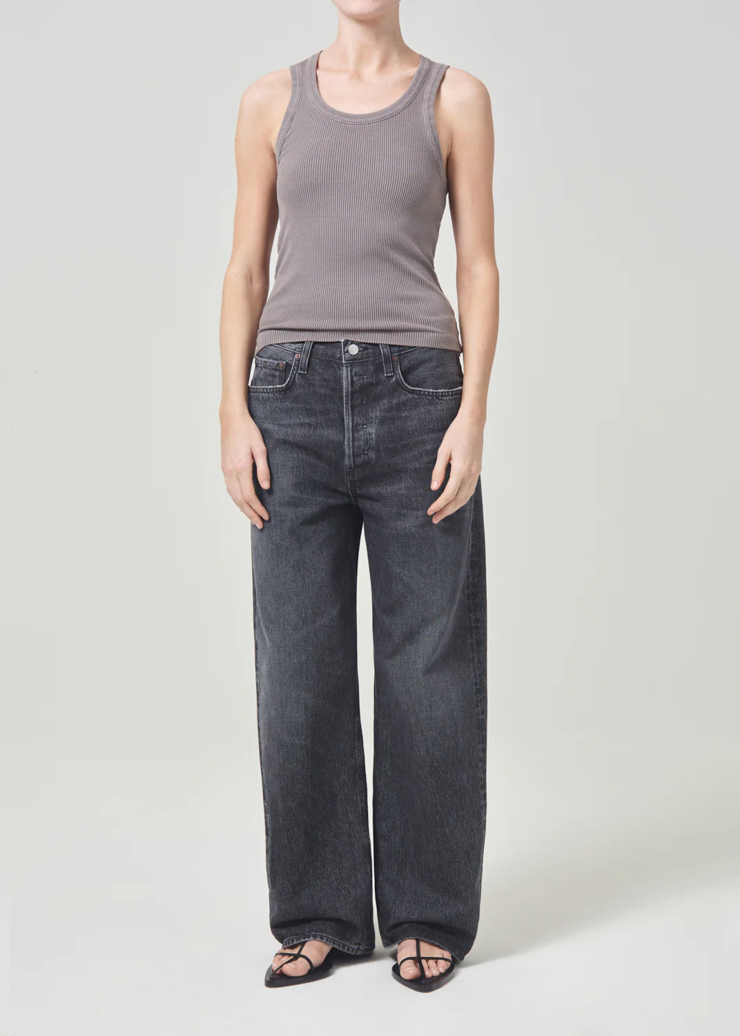 Individual wearing a grey sleeveless top, black AGOLDE Low Slung Baggy 30.5" - Ditch jeans, and black sandals, standing against a plain background.