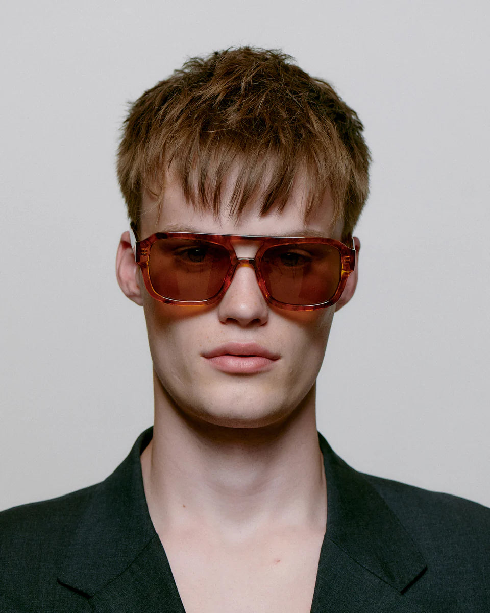A young person with short, tousled brown hair wears A. Kjærbede's Kaya Sunglasses - Havana, featuring large square frames and UV 400 protection, paired with a dark jacket and facing the camera against a light background.