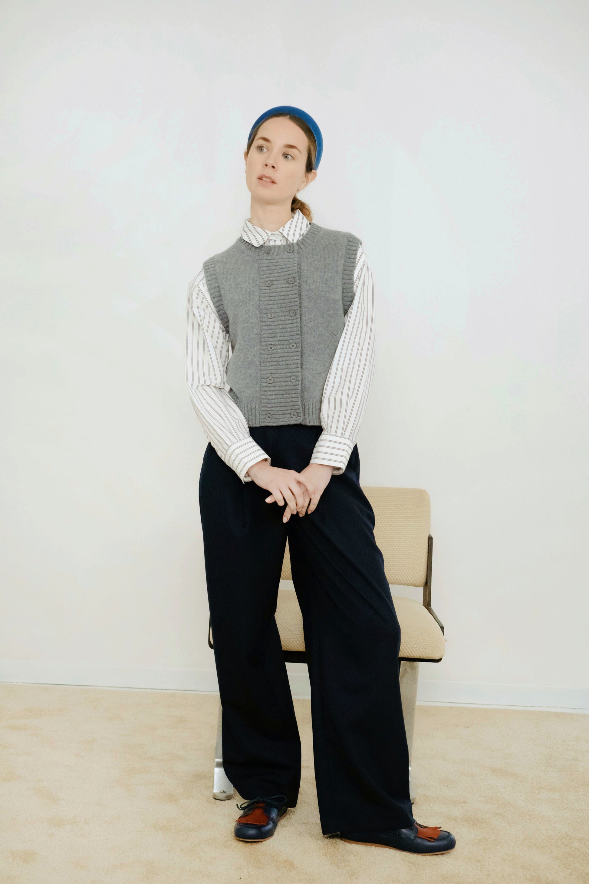 A person stands indoors by a chair, wearing the Tiny Big Sister Buttoned Vest in Dark Grey over a striped shirt, paired with wide-leg black pants and a blue headband.