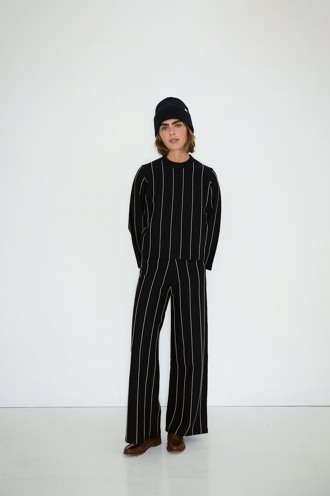 A person stands against a white background wearing the Tiny Big Sister Chloe Striped Sweater in navy, paired with black pants featuring vertical white stripes and brown shoes.