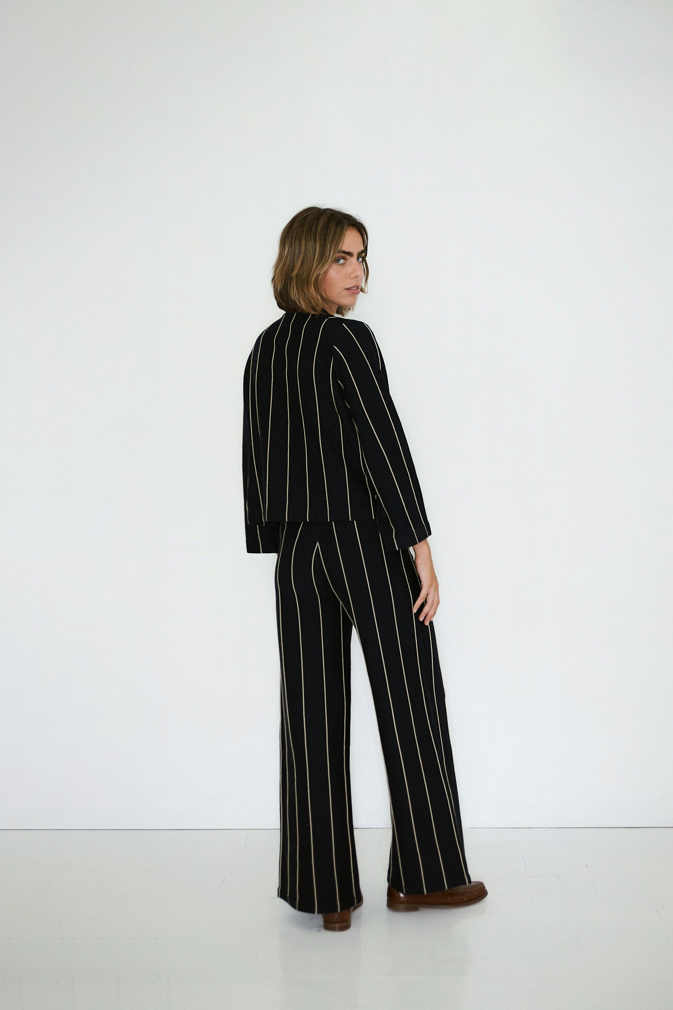 Person stands before a white wall, looking over their shoulder, wearing the Tiny Big Sister Chloe Striped Sweater in navy with vertical white stripes, paired with black suit pants and brown shoes.