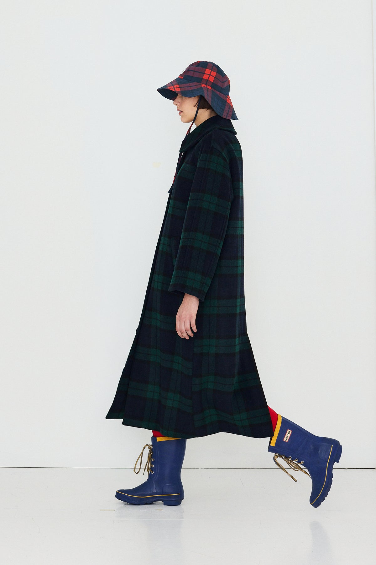 A model in profile wearing a long dark green and black plaid coat, a matching plaid bucket hat, and blue rain boots against a plain white studio background.