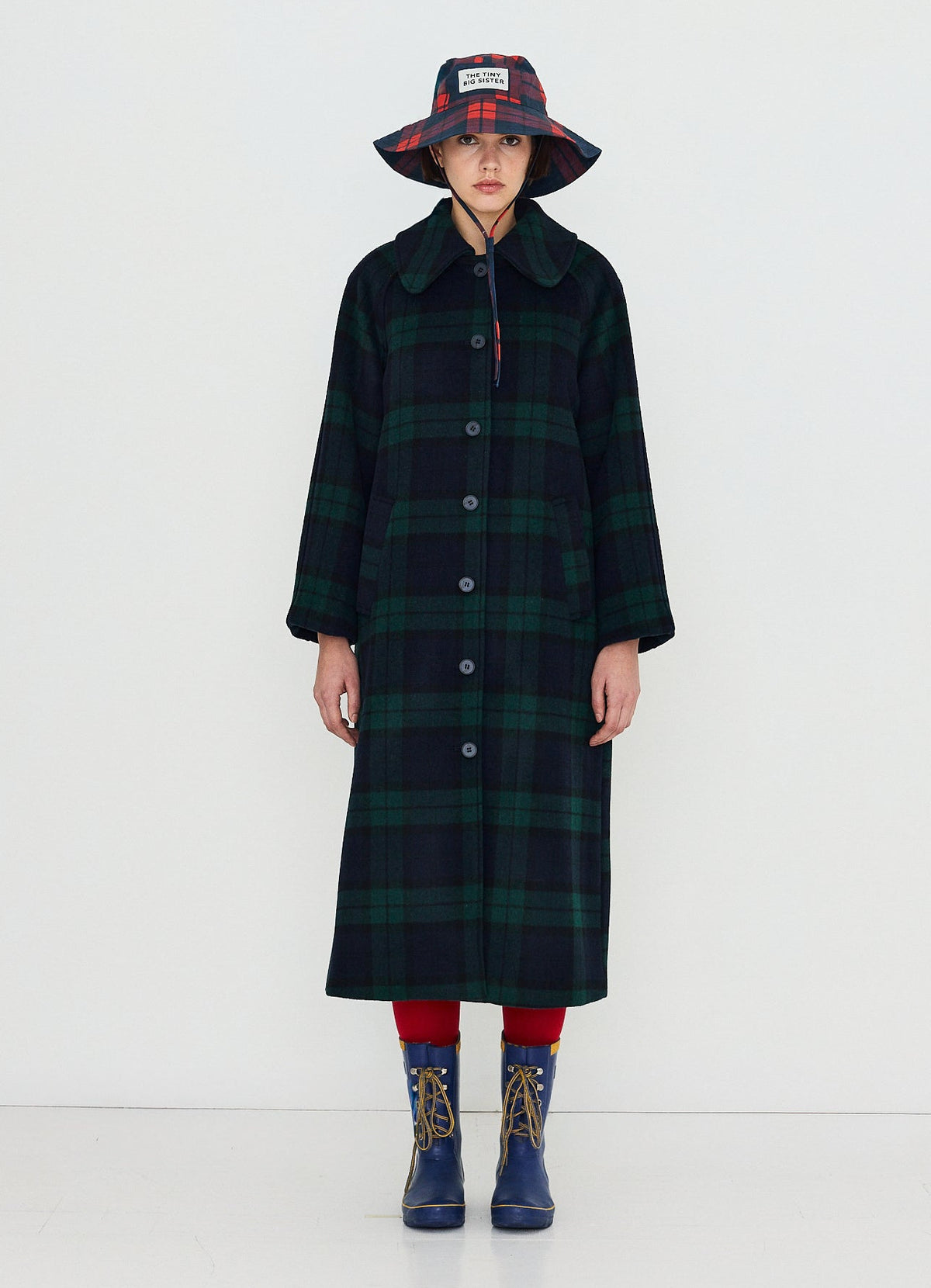 A model stands against a plain white studio background wearing a long dark green and black plaid coat, a matching plaid bucket hat, red tights, and blue rain boots.
