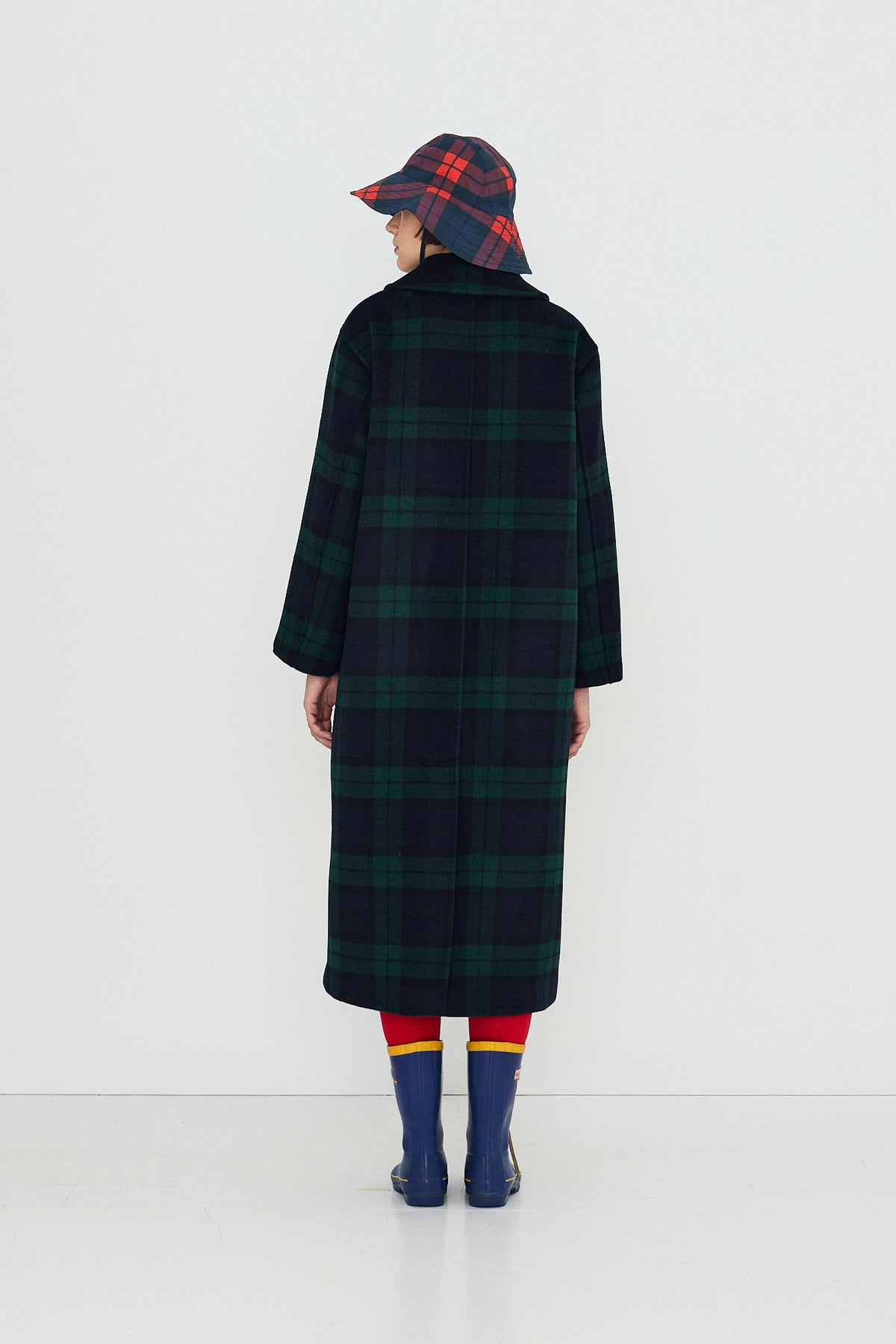 A model shown from the back wearing a long dark green and black plaid coat, a matching plaid bucket hat, red and yellow striped tights, and blue rain boots against a plain white studio background.