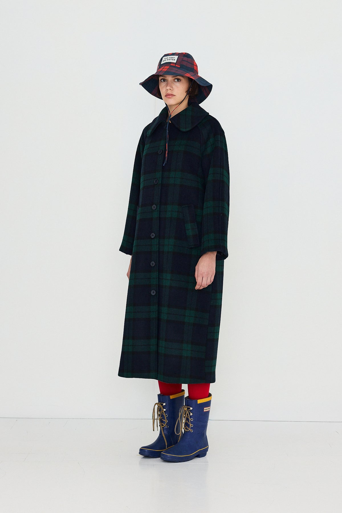 A model stands against a plain white studio background wearing a long dark green and black plaid coat, a red and black bucket hat, red socks, and blue rain boots.