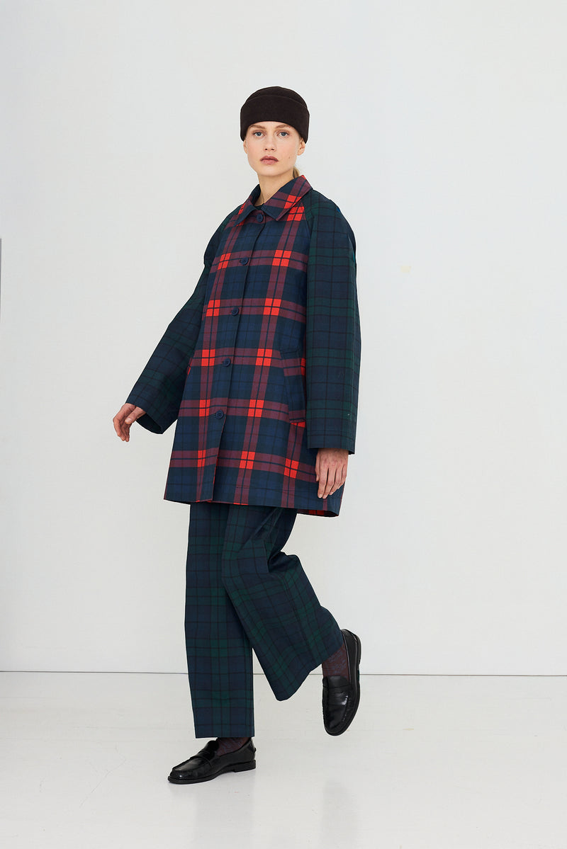 A person dressed in the Check Hood Trench Coat by Tiny Big Sister, layered over a plaid jacket and matching pants, accessorized with a black hat and black shoes, stands against a plain white background for a modern look.