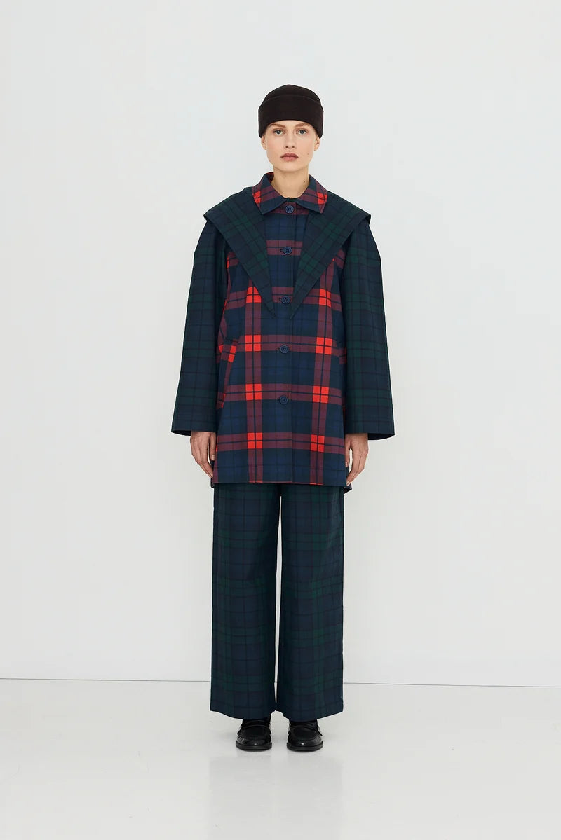 A person stands against a white background wearing a large-collared red plaid shirt, green plaid pants, black shoes, and a dark cap. Layered over the ensemble is the Tiny Big Sister Check Hood Trench Coat—a water-resistant cotton trench that adds both functionality and style to the striking look.