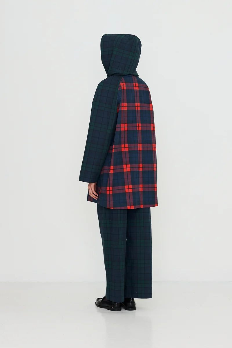A person stands against a plain background wearing the Tiny Big Sister Check Hood Trench Coat—an eye-catching water-resistant cotton trench featuring dark green and blue plaid, a bold red and blue plaid panel on the back, and black shoes for a stylish yet functional look.