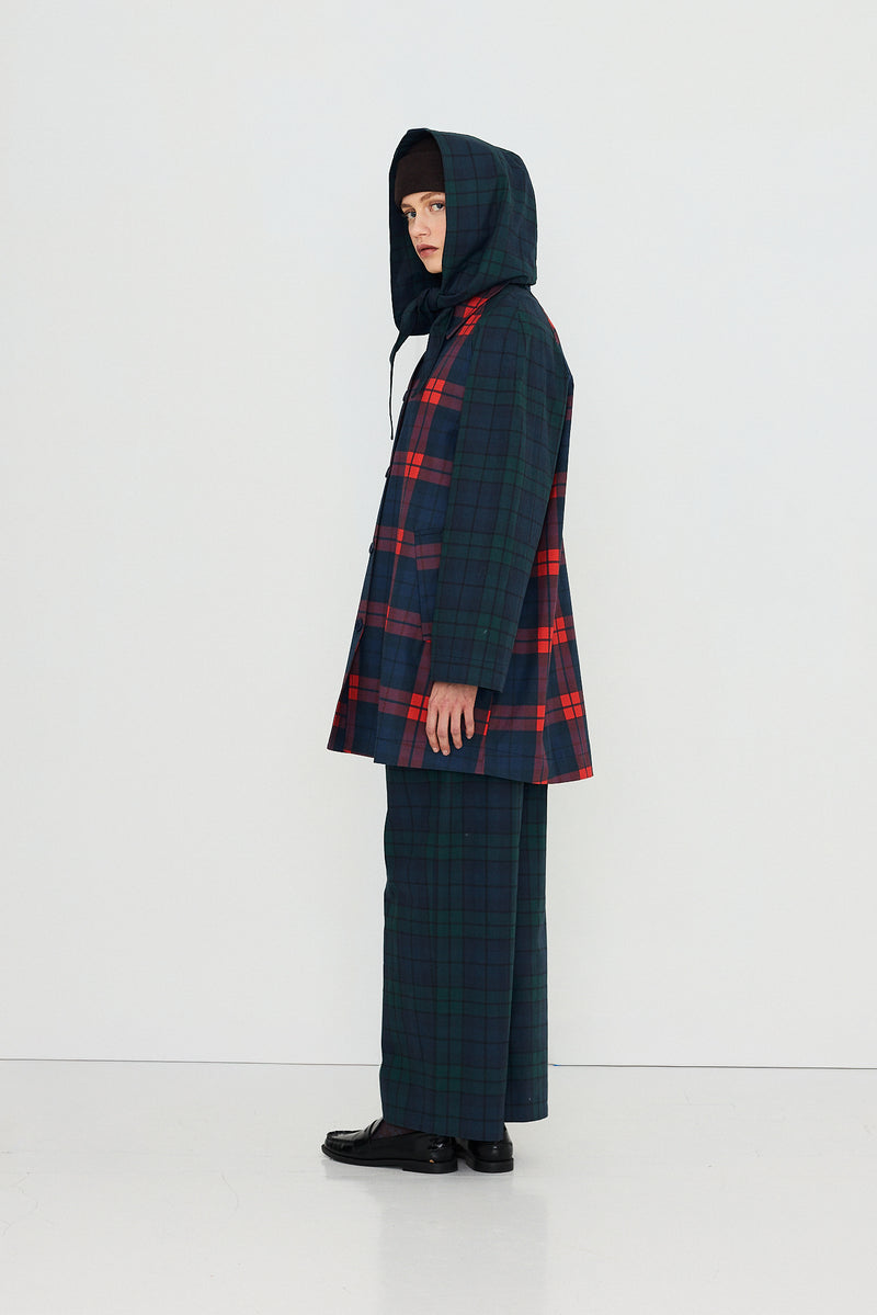 A person stands sideways against a plain white background, wearing the Tiny Big Sister Check Hood Trench Coat in an oversized multicolored plaid pattern, paired with matching wide-leg pants and black shoes.