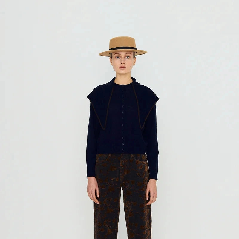 A person stands against a plain background, wearing Tiny Big Sister’s Scarf Neck Lara Cardigan in navy—a merino wool piece featuring shoulder details and a scarf neckline—styled with a tan hat and patterned brown pants.