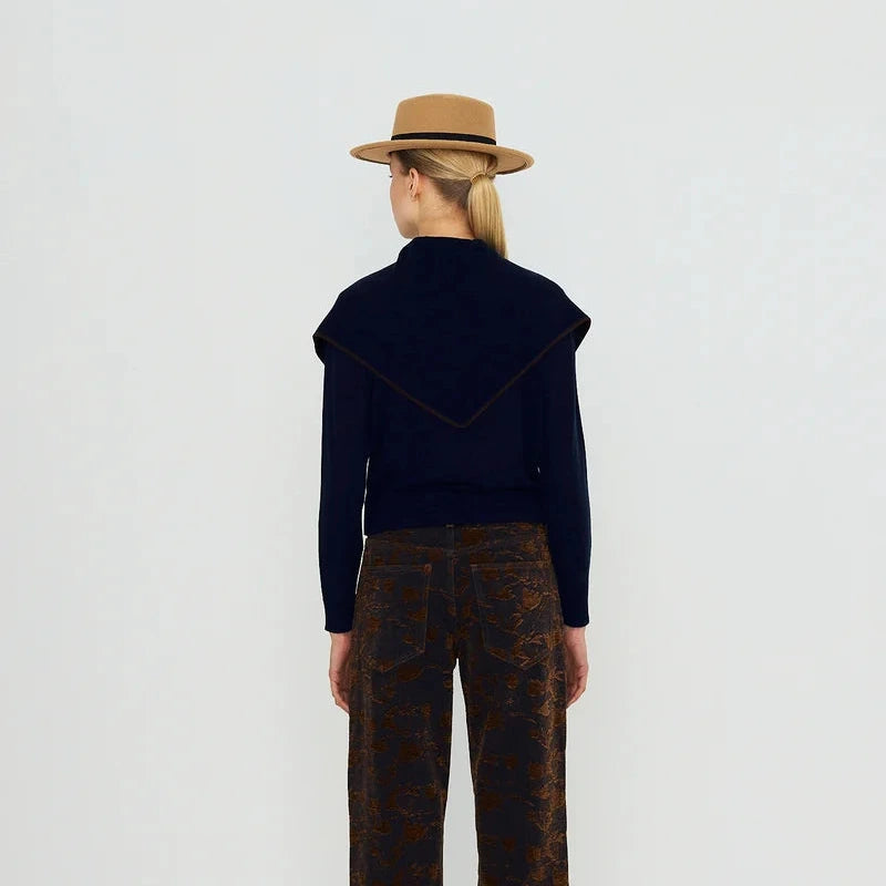 A model viewed from the back wearing a tan fedora hat, a dark navy long-sleeve top with a wide collar, and brown wide-leg trousers with a subtle pattern.