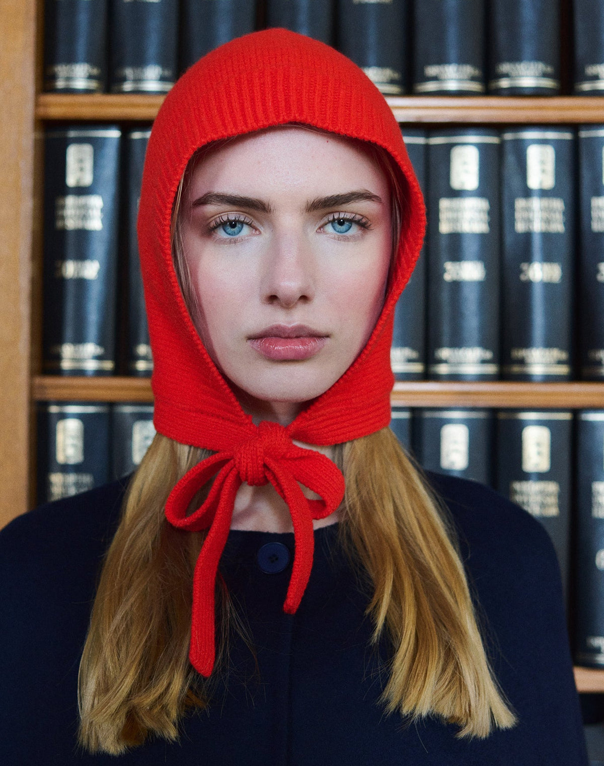 A person with long blonde hair stands before shelves of black hardcover books, wearing the Tiny Big Sister Merino Bonnet - Summer Red with a bow; the scene evokes a minimalist silhouette.
