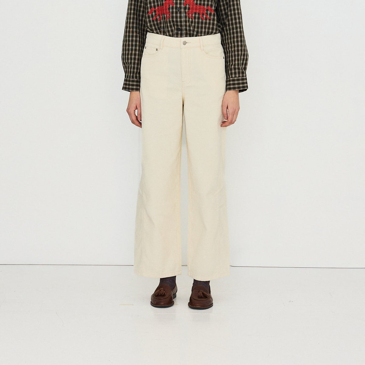 A model stands against a plain white studio background wearing a tan wide-brim hat, a black and brown checkered shirt with red embroidered details, cream wide-leg trousers, and dark brown shoes.