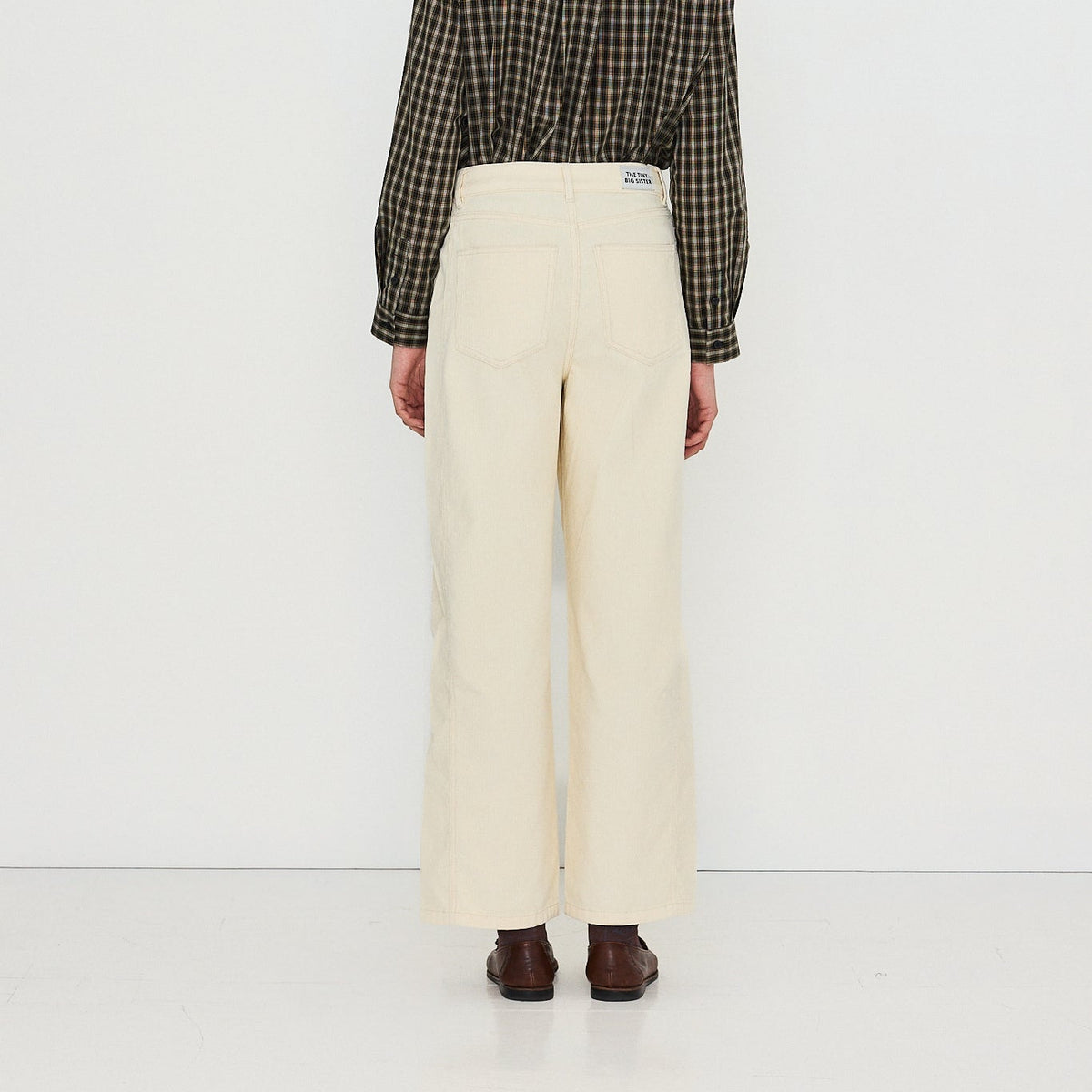 A model viewed from the back wearing a tan fedora hat, a dark green and black checkered long-sleeve shirt, and high-waisted cream wide-leg trousers, standing against a plain white studio background.