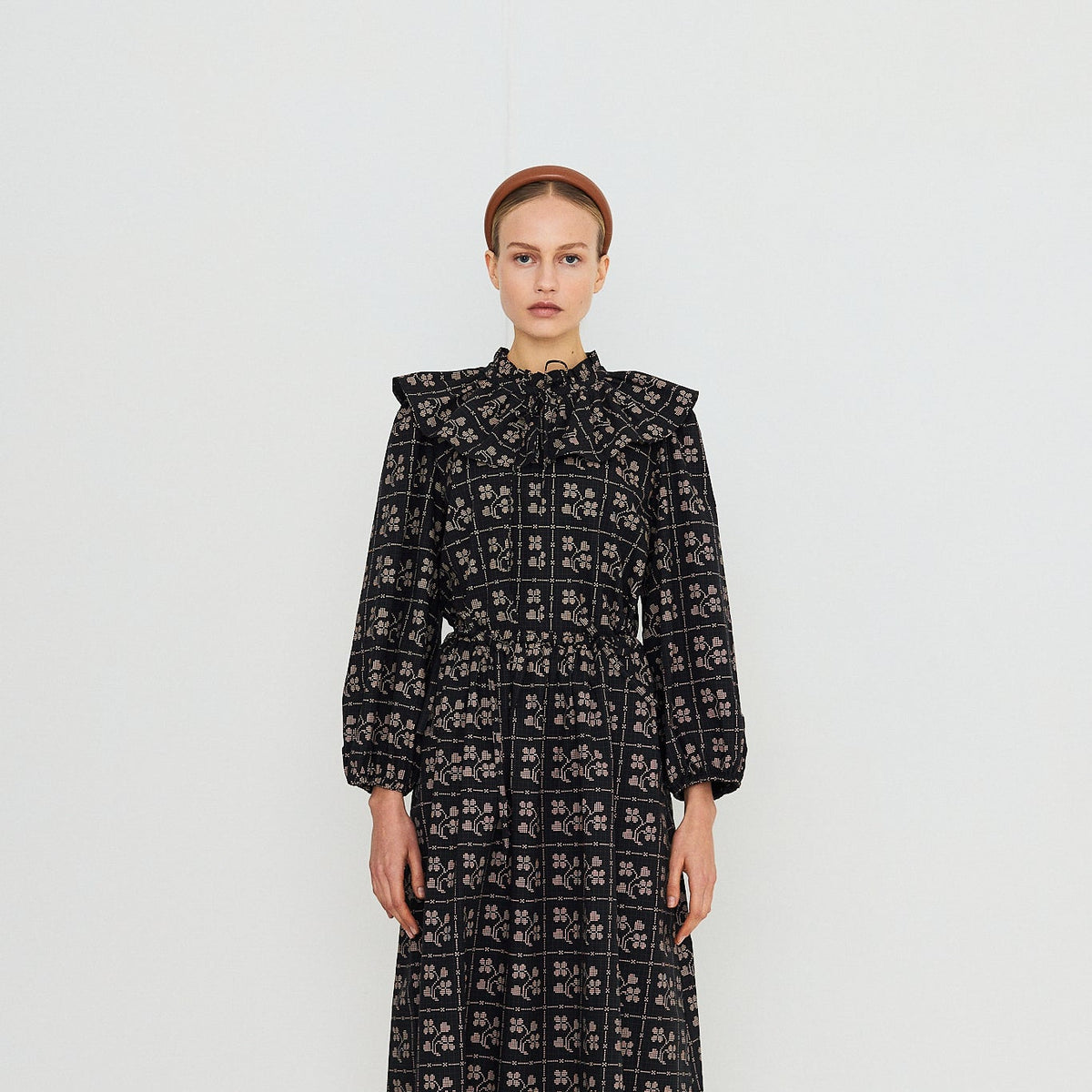 A model stands against a plain white studio background wearing a long-sleeved midi dress with a high ruffled collar and an all-over black and white geometric pattern. She is also wearing red socks and dark brown shoes.