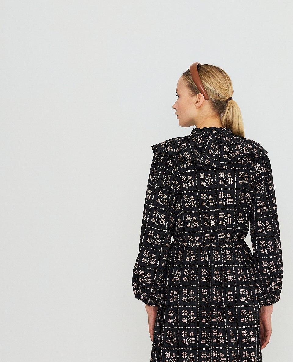 A woman stands with her back to the camera wearing a long-sleeved midi dress with a black and white geometric pattern, paired with red socks and brown shoes, against a plain white studio background.