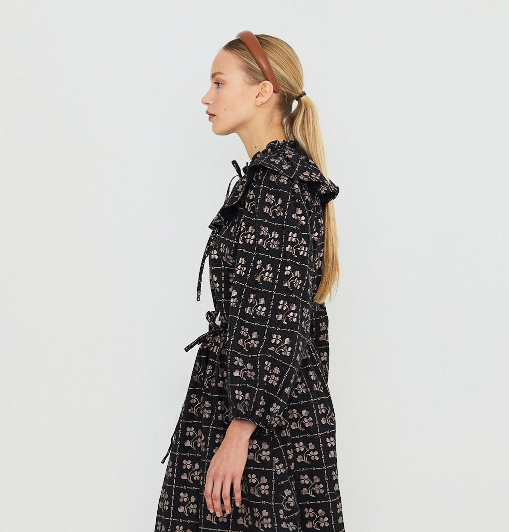 A woman in profile with long blonde hair styled in a low ponytail, wearing a brown headband and the Tiny Big Sister Dots Flowers Puff Sleeves Blouse in Navy—featuring floral designs and a subtle checked dot pattern—stands against a plain, light background.