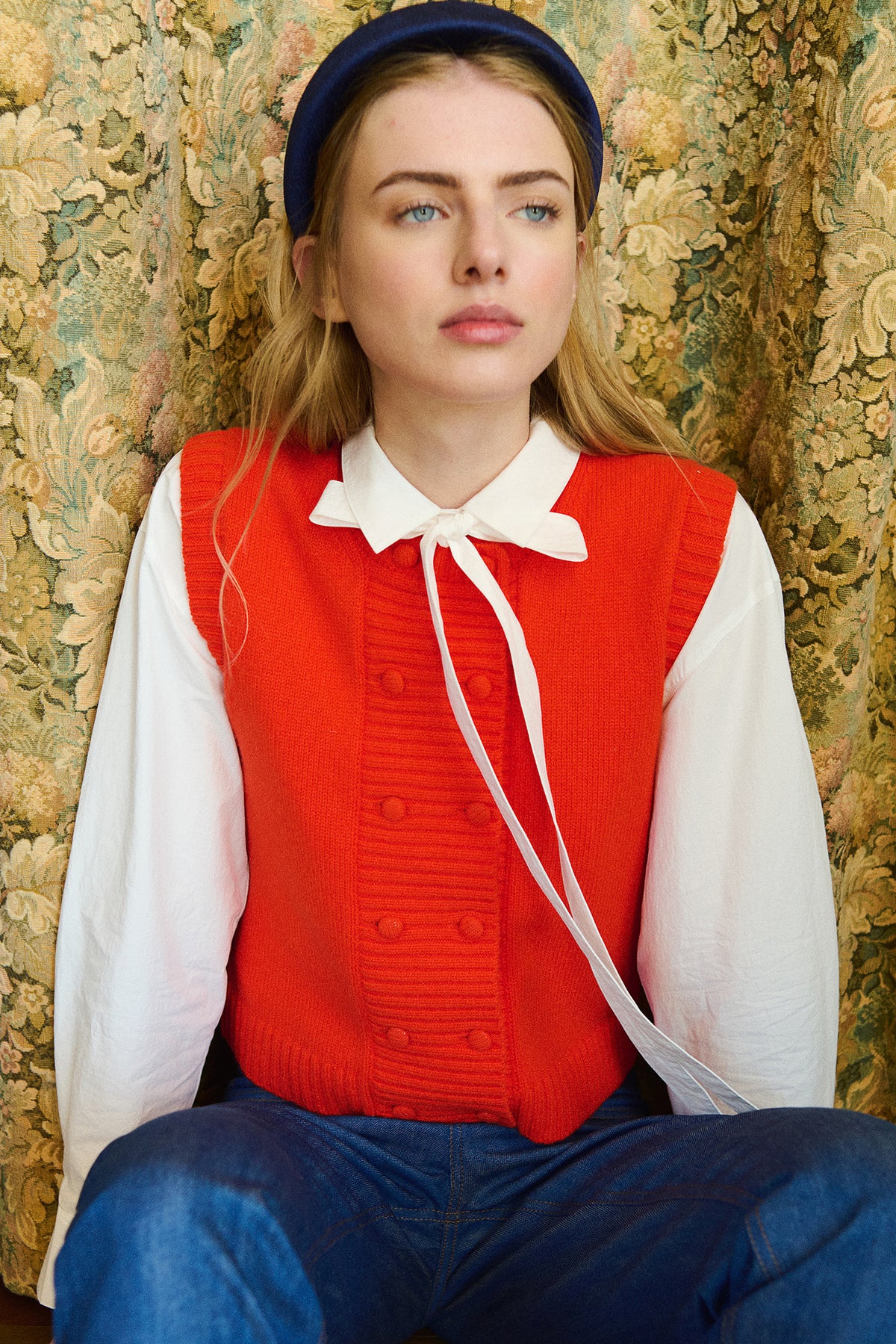 A young woman wearing a bright red ribbed knit vest over a white long-sleeve collared shirt, paired with blue jeans and a navy blue beret, seated against a vintage floral-patterned wallpaper background.