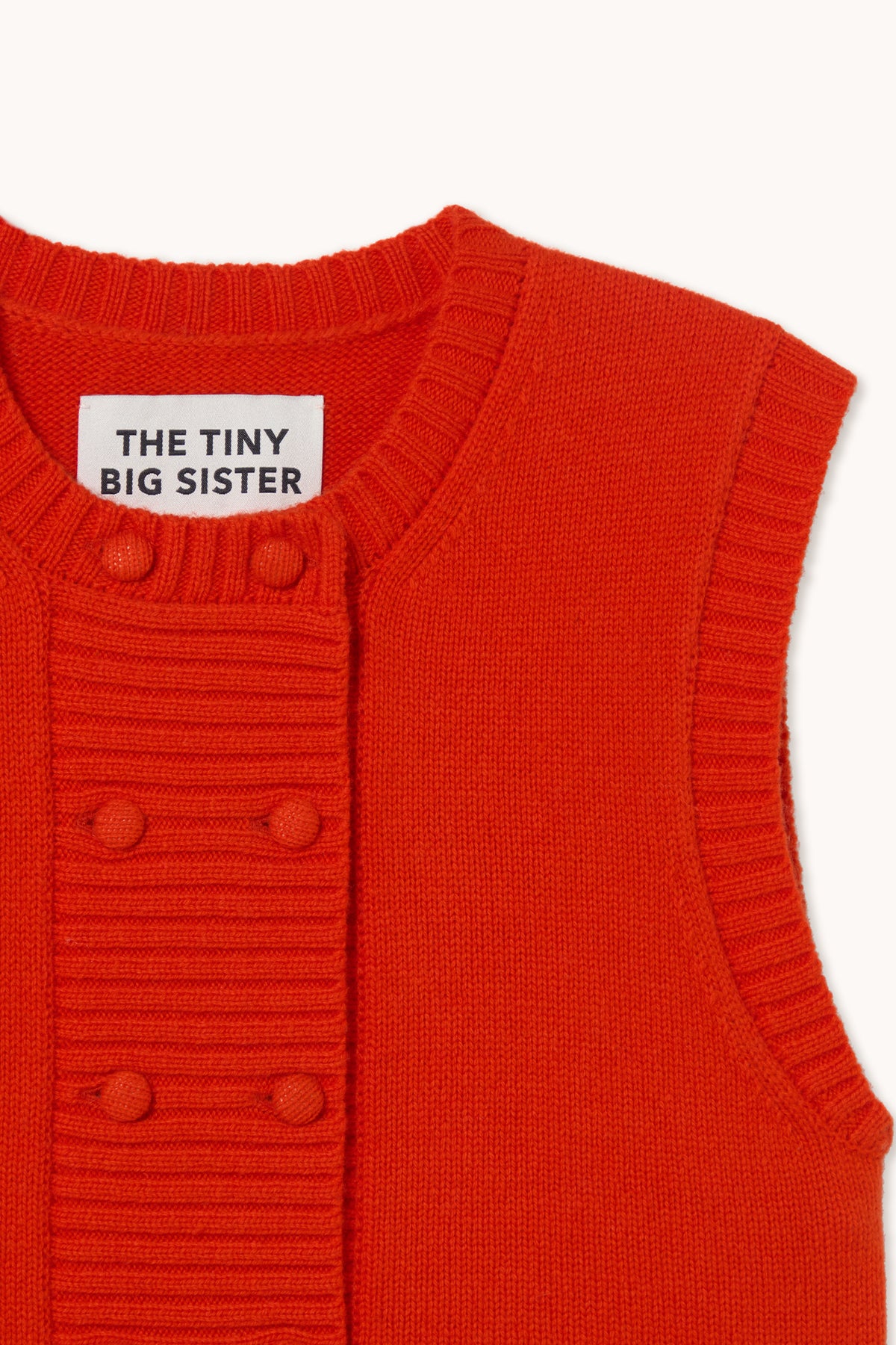 Close-up of a bright red ribbed knit vest with a white neck label reading 'THE TINY BIG SISTER' and three matching red buttons on the front.