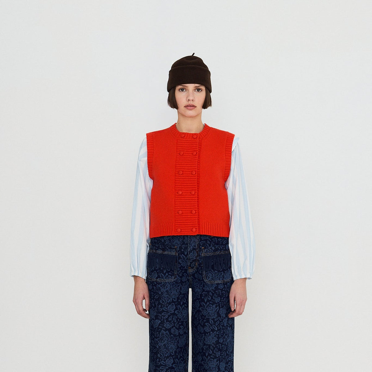 A model stands against a plain white studio background wearing a bright red sleeveless vest over a white long-sleeve shirt, paired with dark blue patterned wide-leg trousers and brown shoes.