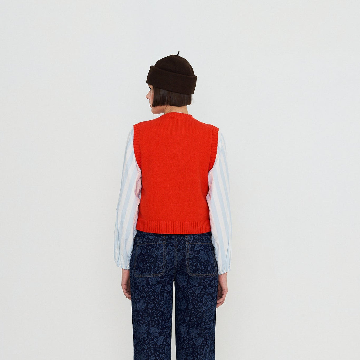 A model viewed from the back wearing a bright red knit sweater vest over a white long-sleeve shirt, paired with dark blue patterned wide-leg trousers and a dark brown beret.