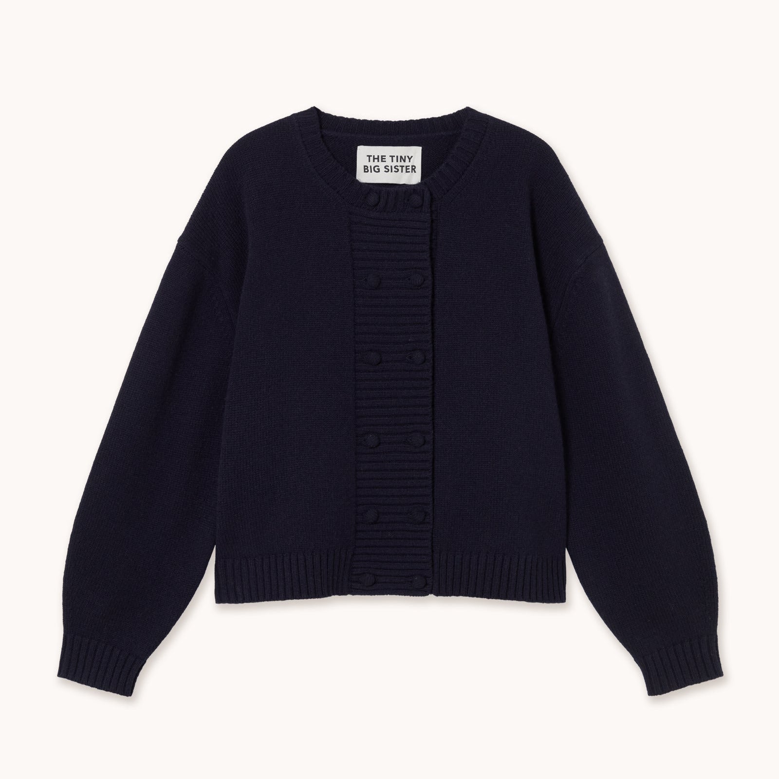 A dark navy blue knit cardigan sweater with a central ribbed button placket and long sleeves, laid flat against a light cream background. The brand label reads tiny big sister.