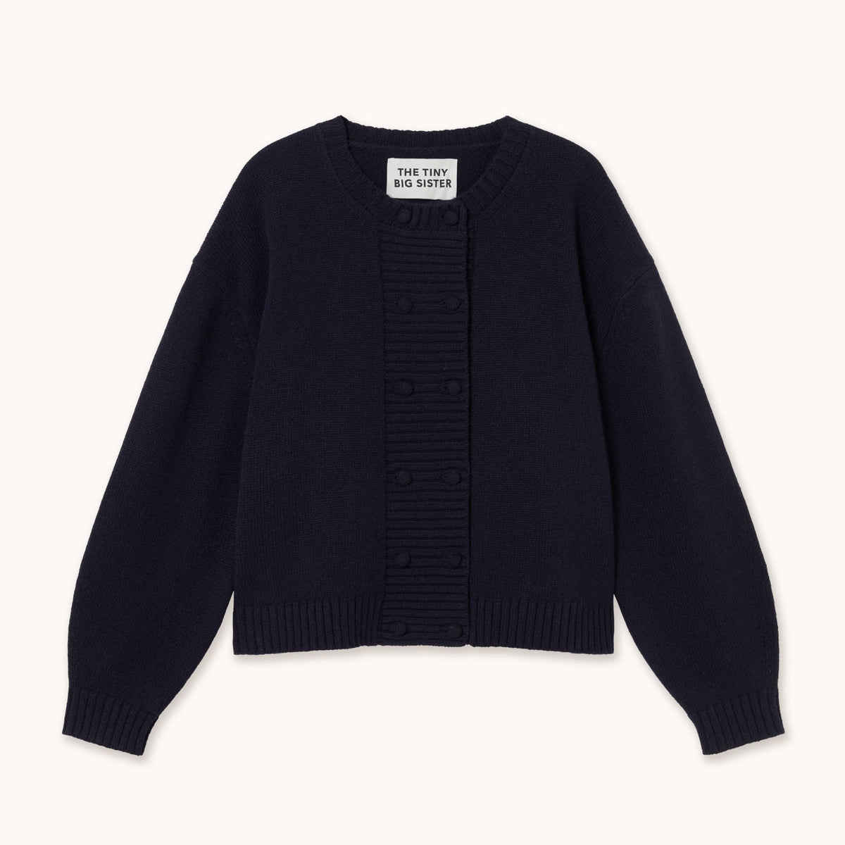 A dark navy blue knit cardigan sweater with a central ribbed button placket and long sleeves, laid flat against a light cream background. The brand label reads tiny big sister.