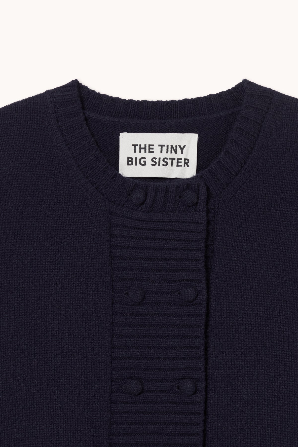 Close-up of a navy blue ribbed knit cardigan sweater with a white neck label reading 'THE TINY BIG SISTER'.