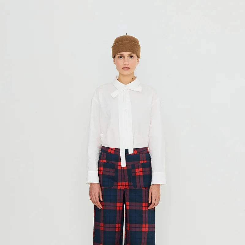 A model stands against a white studio background wearing a white long-sleeve blouse with a bow collar, wide-leg navy and red plaid trousers, a brown beret, and red shoes.