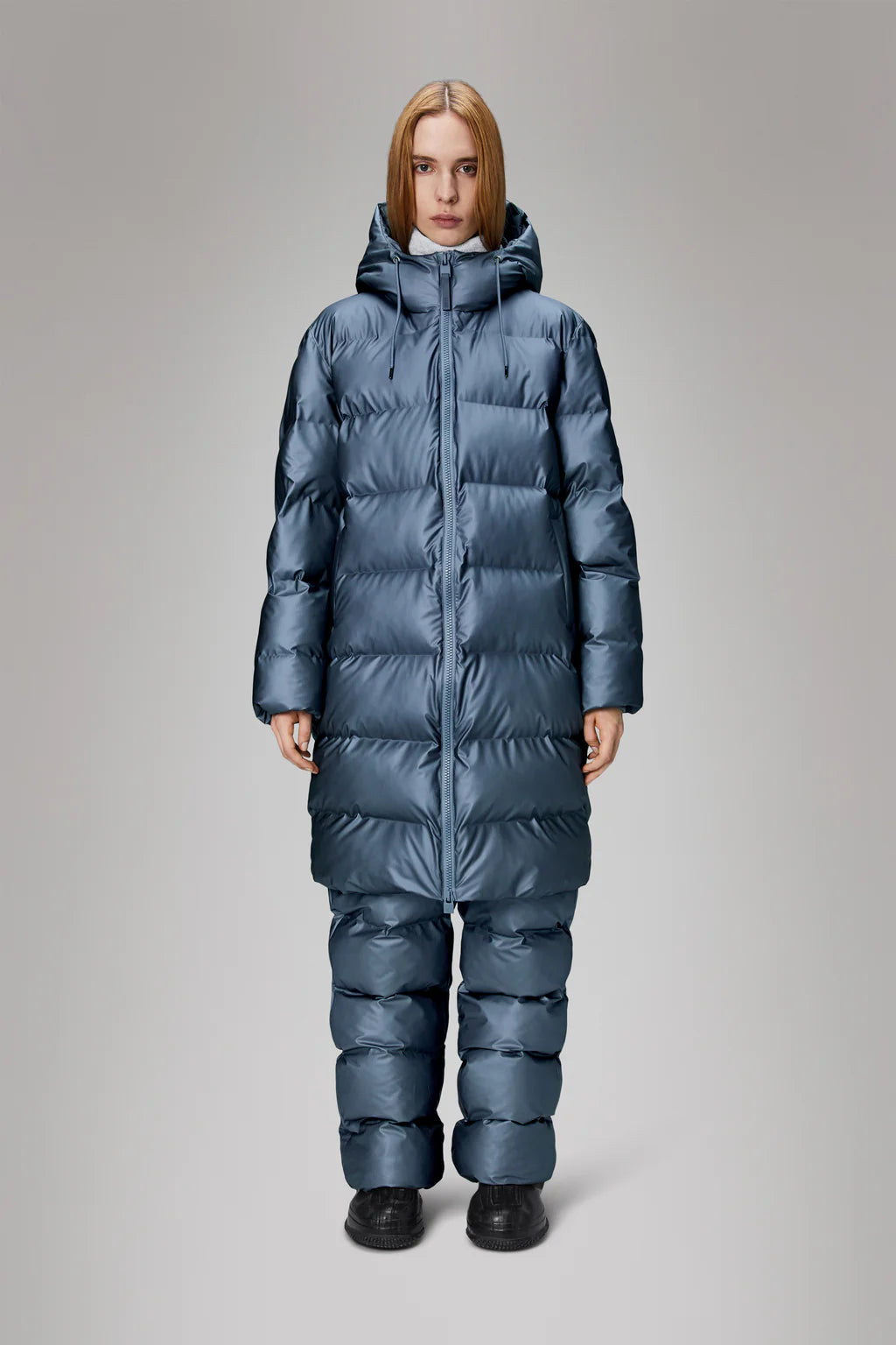 A person wearing an Alta Long Puffer Jacket by Rains in a blue color, featuring breathable fabric and featherless chamber technology, stands against a neutral background.