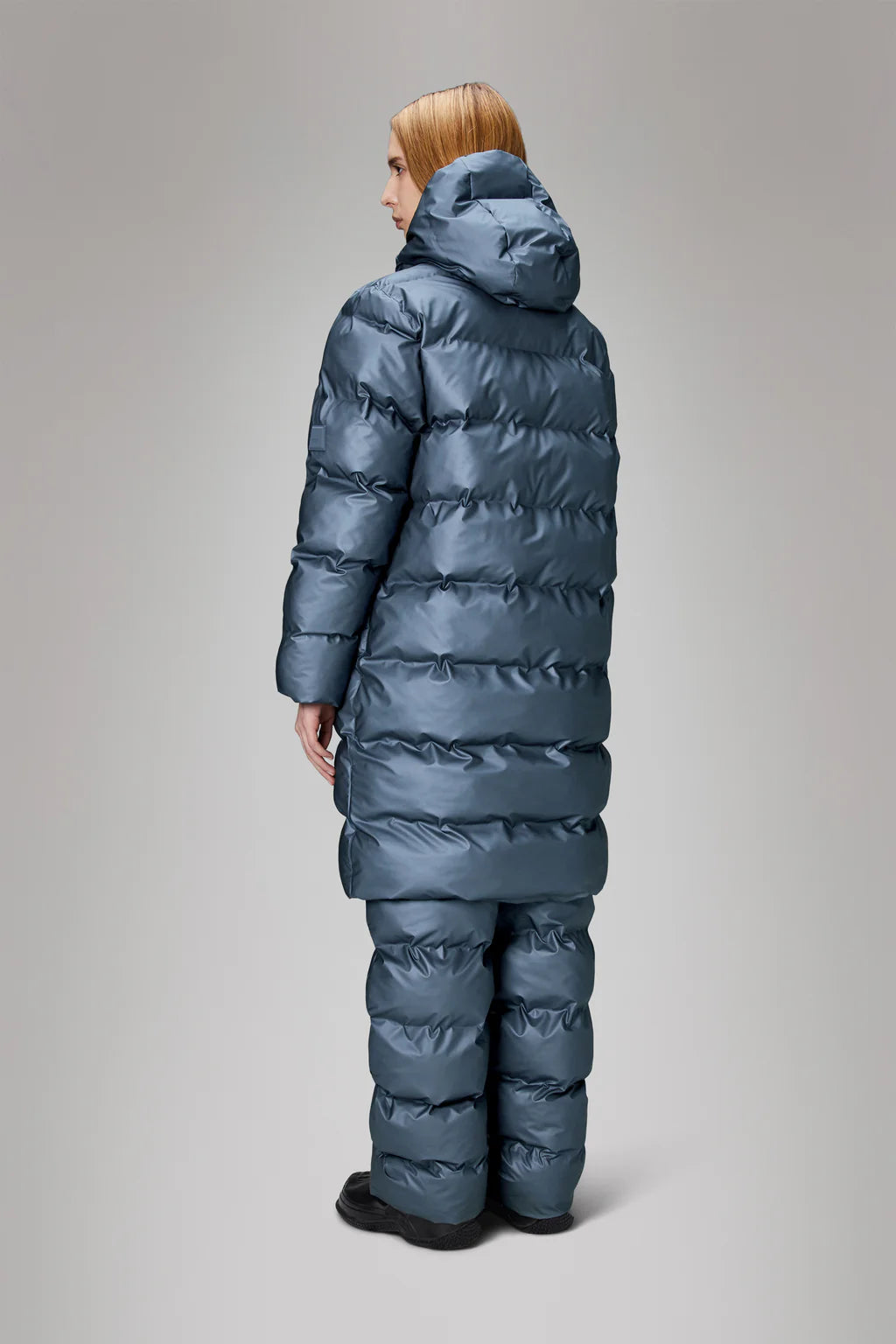 Person wearing the Alta Long Puffer Jacket by Rains, a blue, puffy, full-body winter coat with a hood, seen from the back against a plain background. The jacket features featherless chamber technology for warmth and comfort during cold seasons.