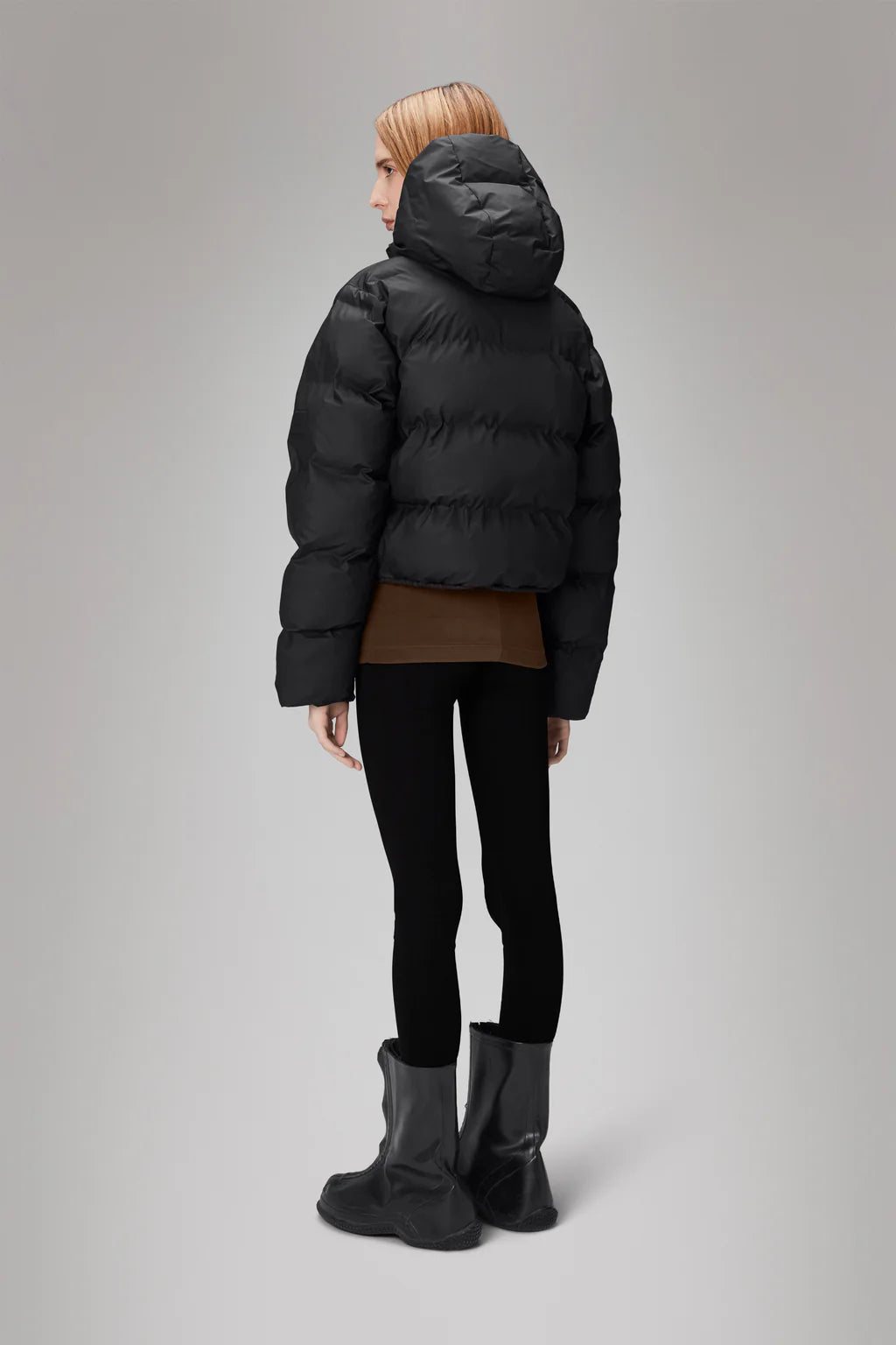 A person is seen from behind wearing the sleek Alta Short Puffer Jacket by Rains, paired with black leggings and black boots, standing against a plain gray background. This stylish outfit not only keeps them warm but also offers the practical benefits of a winter jacket.