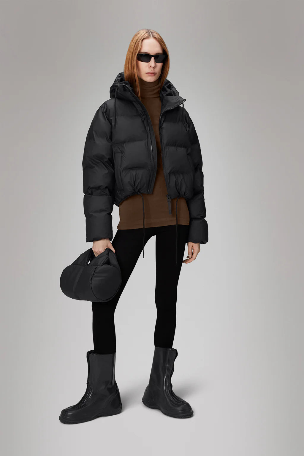 A person wearing an Alta Short Puffer Jacket by Rains, a brown turtleneck, black leggings, black boots, and sunglasses holds a black handbag while standing against a gray background.