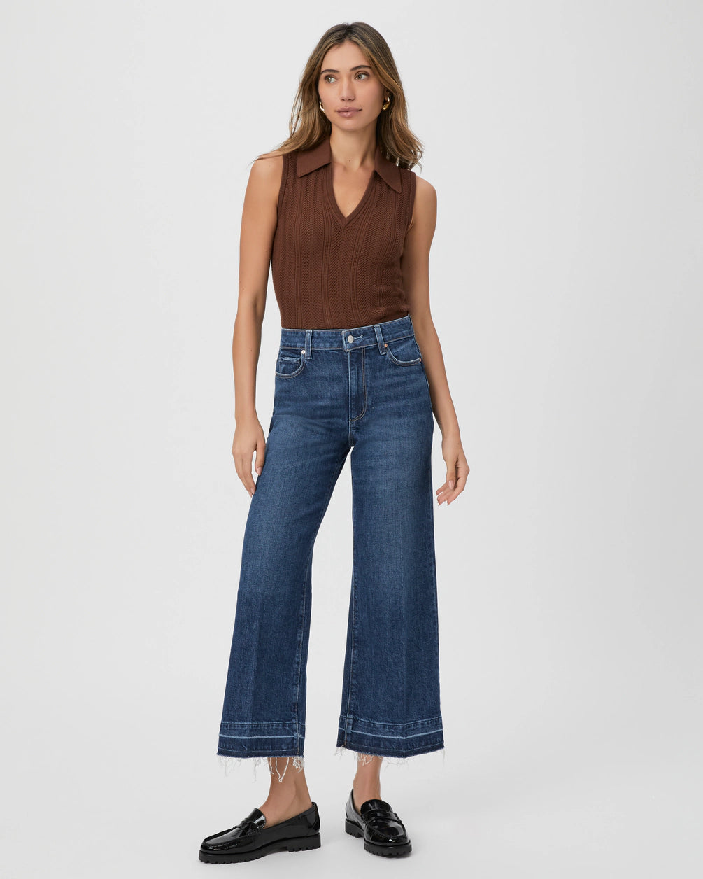 A woman wearing a sleeveless brown top and Paige's Anessa Wide Leg - Alana vintage-inspired, ultra high-waisted wide-leg blue jeans stands with her left hand on her hip. She is also wearing black loafers. The background is white.