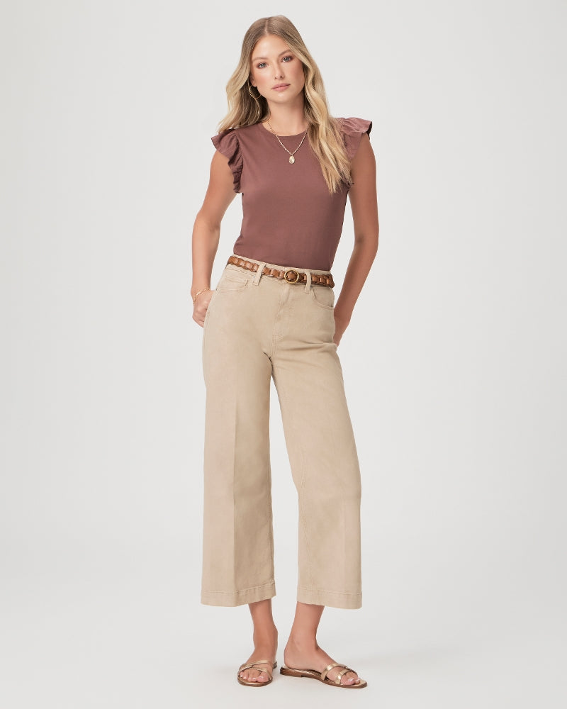 A woman stands wearing Paige's Anessa Wide Leg - Vintage Soft Sand pants, a brown ruffled-sleeve top, a belt, and sandals, posing against a white background.