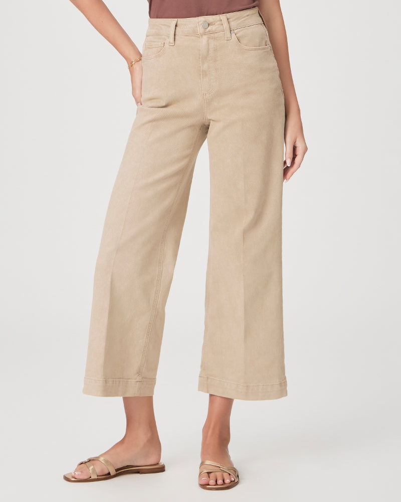 Woman wearing beige high-waisted Paige Anessa Wide Leg - Vintage Soft Sand pants and gold sandals, standing with one hand in pocket against a white background.