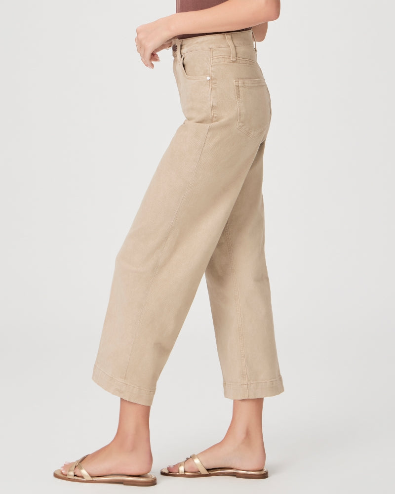 Woman wearing cropped beige high-waisted Anessa Wide Leg - Vintage Soft Sand pants by Paige and gold sandals, standing side-view with hand on waist against a plain background.