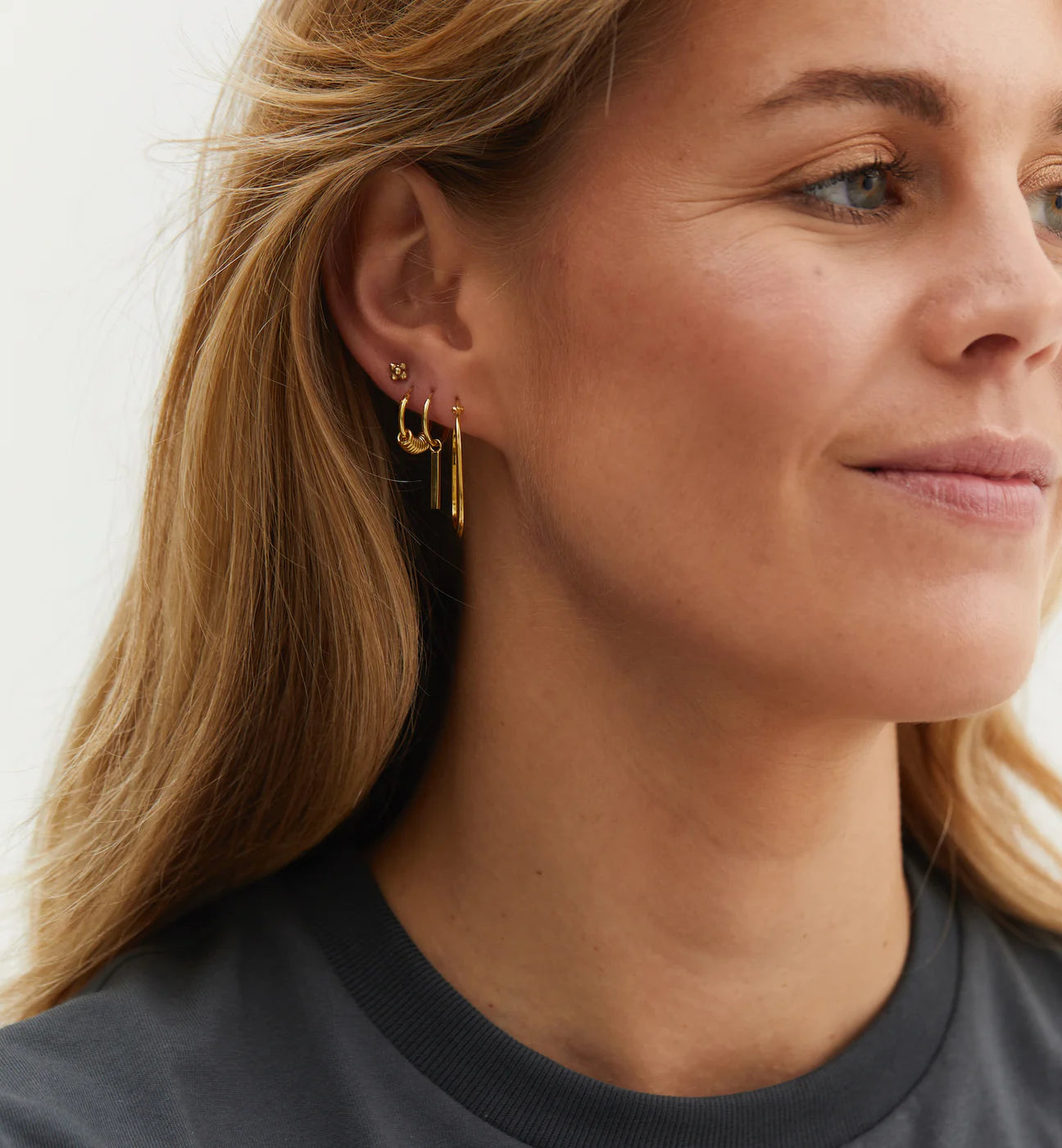 The Anna + Nina Link Hoop Earrings - Goldplated, with their distinct geometric shape, adorn the ears of a woman casually dressed in a t-shirt.