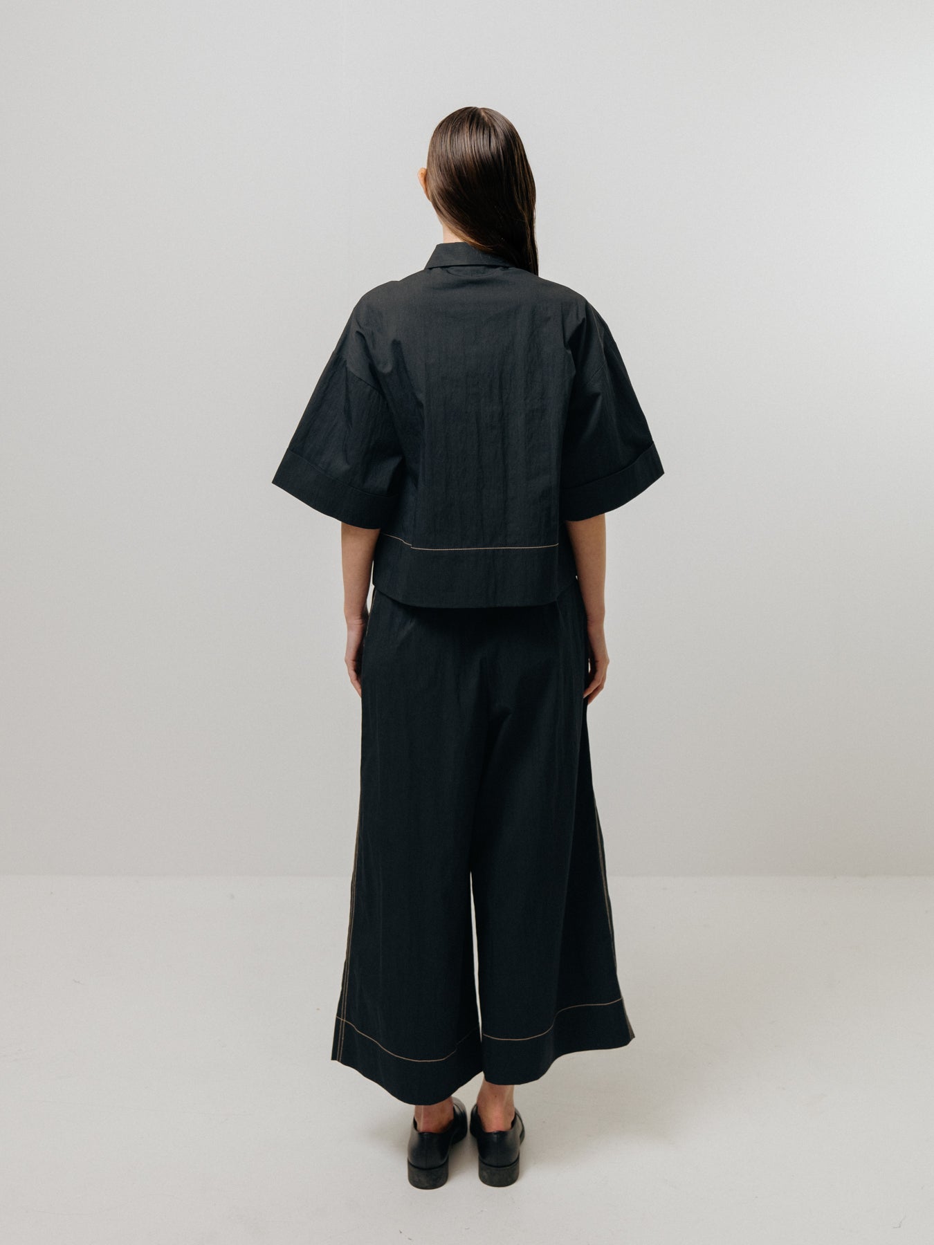 An individual wearing Emin and Paul's Contrast Stitch Shirt - Black faces away in a simple, light-toned room.