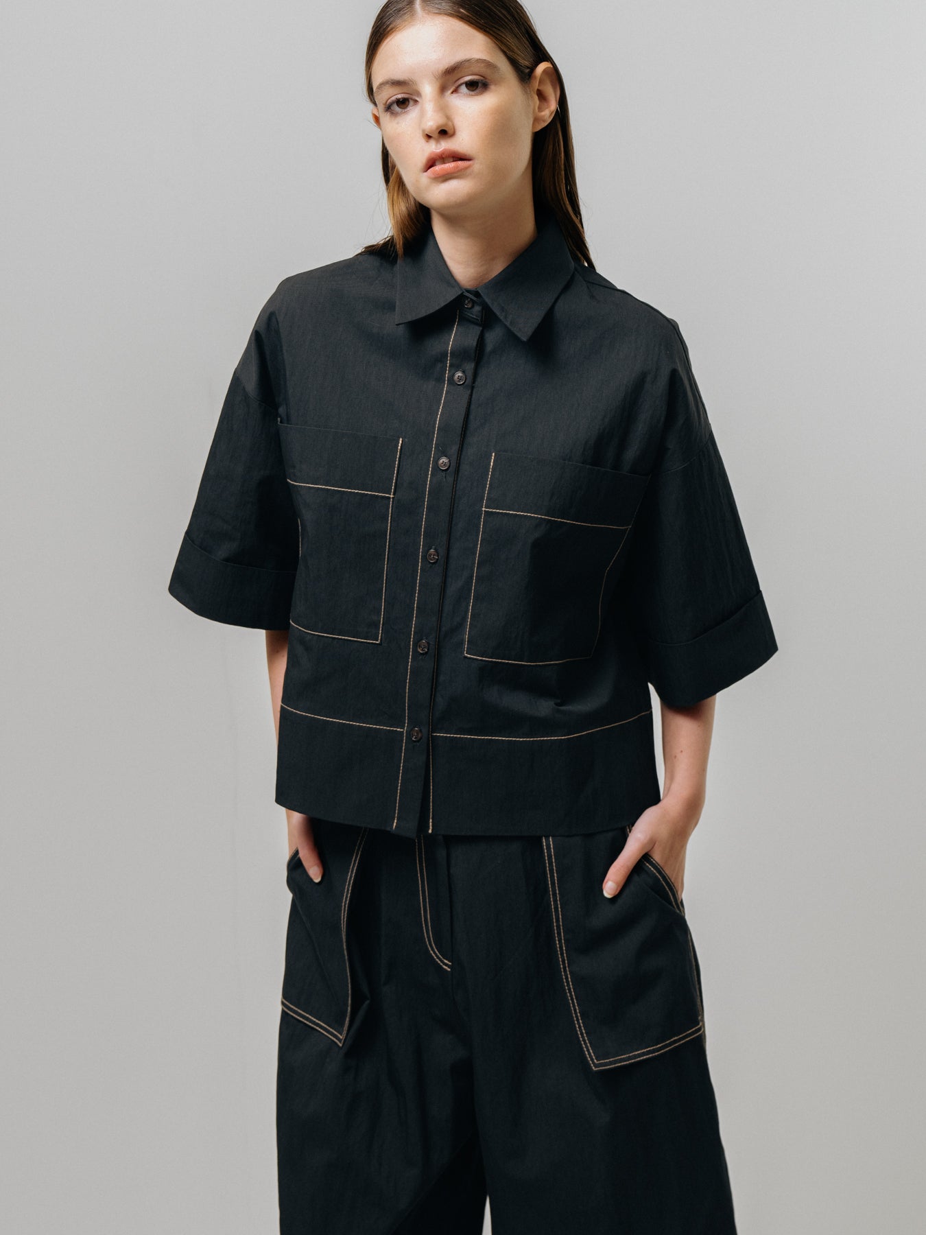 A person confidently poses against a plain background wearing the Emin and Paul Contrast Stitch Shirt - Black, featuring pocket details and contrast stitching, paired with matching wide-leg trousers and hands in pockets.