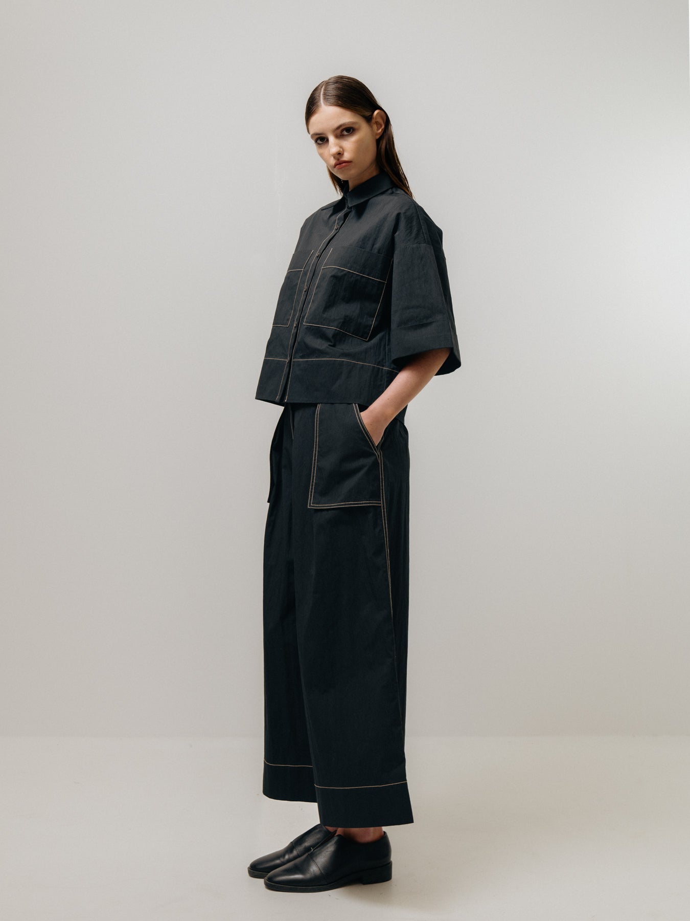 An individual poses against a plain background wearing the Emin and Paul Contrast Stitch Shirt - Black, featuring a short-sleeved jacket with contrast stitching and matching loose-fitting trousers.