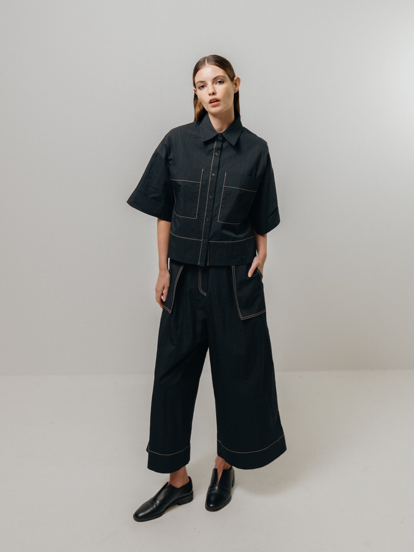 Wearing Emin and Paul's Contrast Stitch Shirt in black, paired with matching wide-legged trousers, a person stands against a plain backdrop.