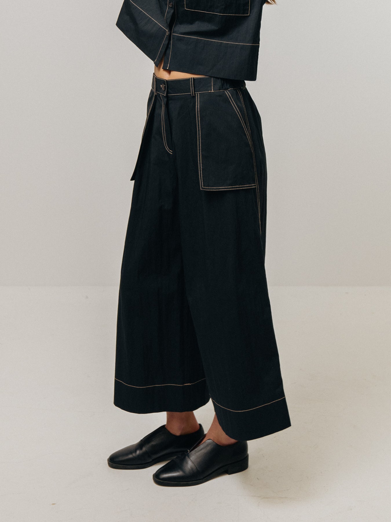 An individual is dressed in a black cropped jacket and Emin and Paul's Wide Leg Trousers - Black with ecru stitching, complemented by black shoes.