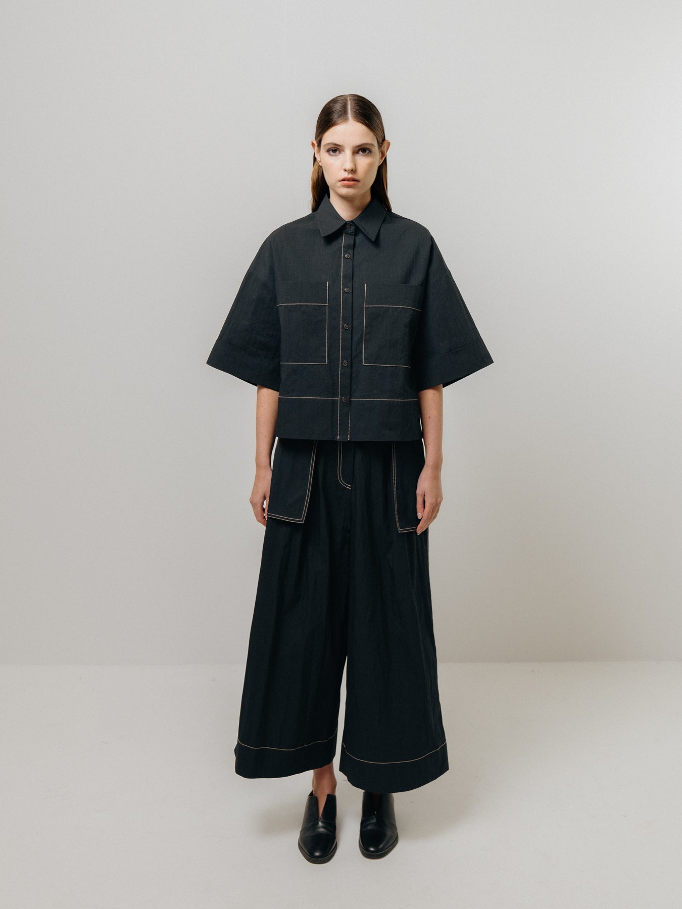 Against a plain backdrop, a person wears Emin and Paul's black wide-leg trousers paired with a loose dark shirt featuring large front pockets. Their straight, shoulder-length hair complements the ensemble, highlighted by ecru stitching detail and finished with black shoes.